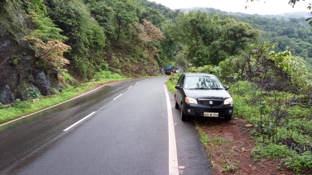 Black Maruti Alto K10 parked to the side of a road slick with recent rain. Forested slopes in the background.